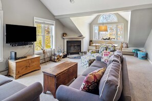 A spacious living room with a fireplace, a television, and a large window with a view of trees. - Okemo Winterplace 2BR Loft – Family Ski Getaway (Ludlow)