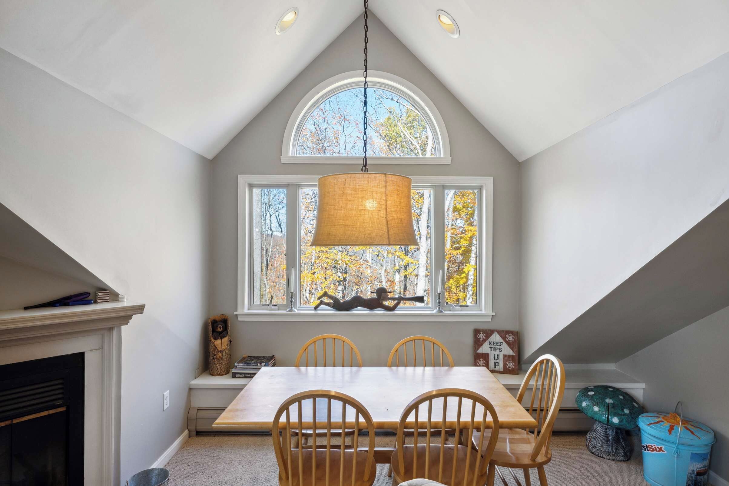 A dining area with a high ceiling, a large window with a view of trees, a hanging light fixture, and a fireplace.