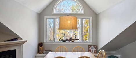A dining area with a high ceiling, a large window with a view of trees, a hanging light fixture, and a fireplace.