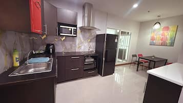 A modern kitchen with dark cabinetry, a stainless steel sink, a microwave, a built-in oven, and a refrigerator. There is a dining area with a table and chairs, and a colorful abstract painting on the wall.