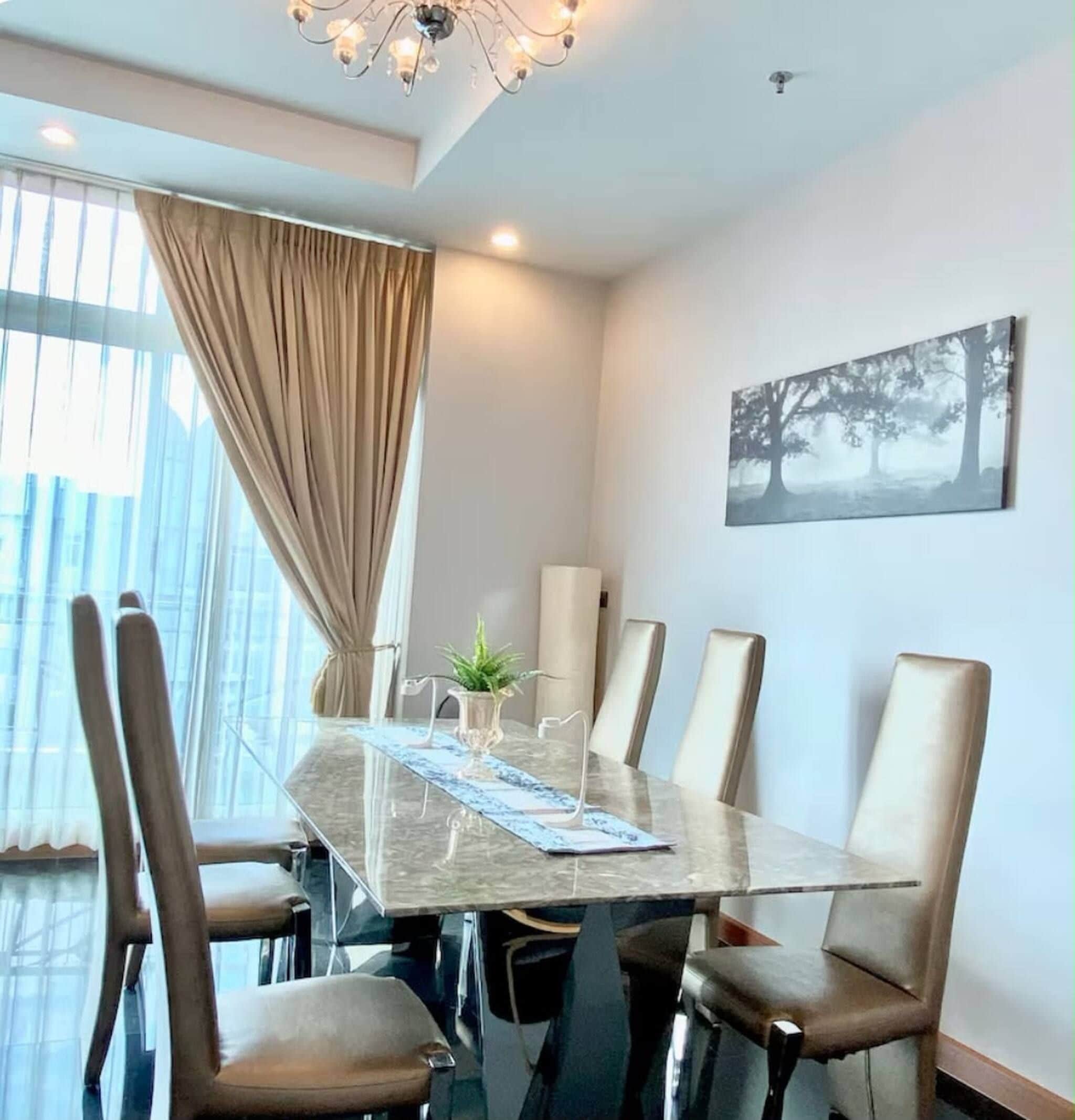 A modern dining room with a glass table, six chairs, a chandelier, a large window with curtains, and a painting on the wall.