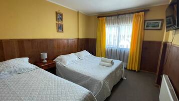 A hotel room with two beds, a bedside table with a lamp, a television, and a window with curtains.
