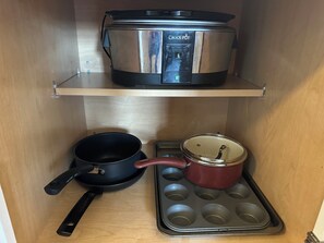 A kitchen cabinet with a stainless steel Crock Pot on the top shelf, a black frying pan with a red handle, and a muffin tray with a red pot on the bottom shelf.
