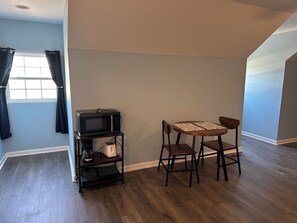 Une pièce avec un plancher en bois, une table entourée de chaises, un four à micro-ondes posé sur une étagère et une fenêtre avec des rideaux.