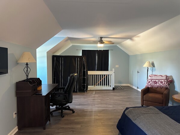 A room with a desk, office chair, and a bed. There is a ceiling fan, a window with curtains, and a small table with a lamp.
