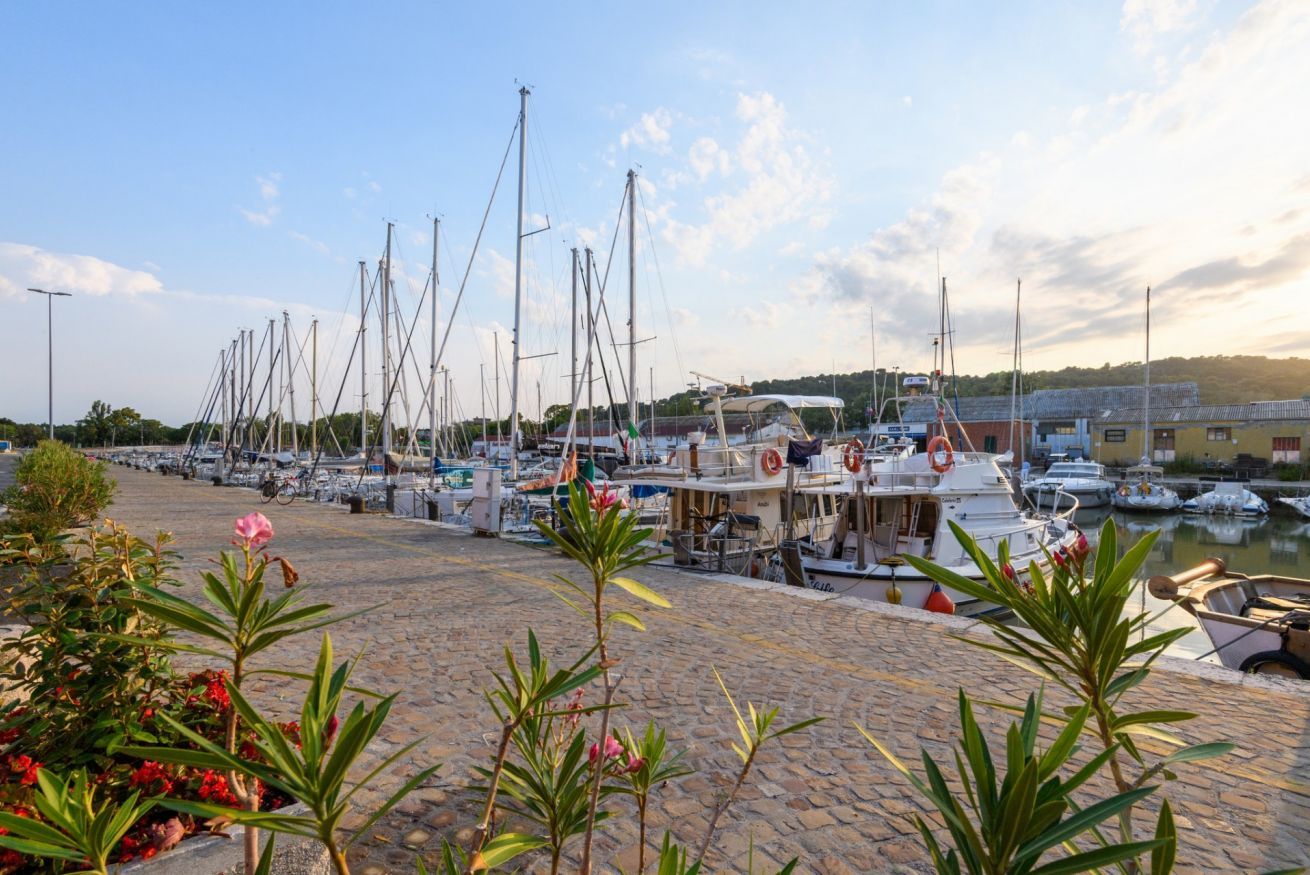 Un port esportiu amb diverses embarcacions amarrades, un passeig de pedra i un edifici amb zona coberta al fons.