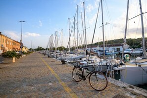 Ein Yachthafen mit vertäuten Booten entlang einer gepflasterten Uferpromenade, ein Fahrrad, das am Gehweg abgestellt ist, und Gebäude im Hintergrund.