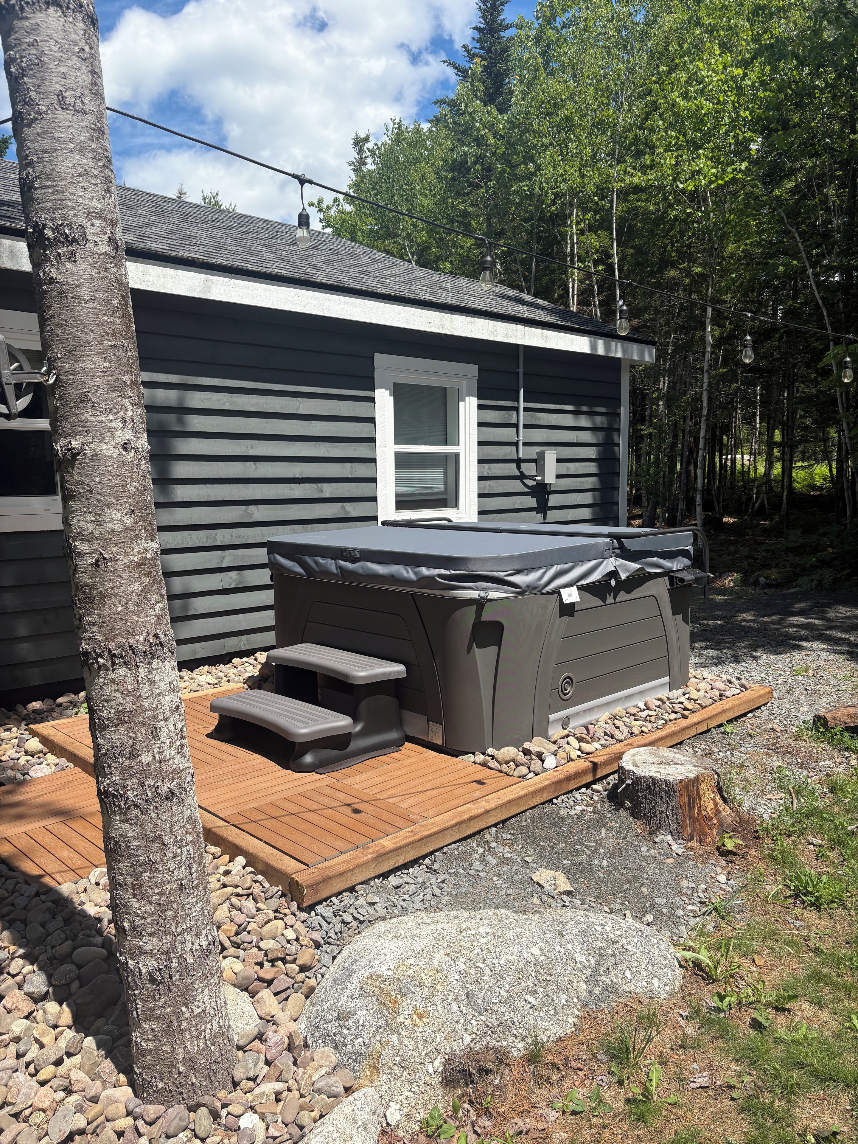 A hot tub with a wooden deck, surrounded by a stone border, next to a house with a window and a chimney.