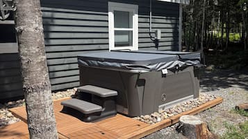 A hot tub with a wooden deck, surrounded by a stone border, next to a house with a window and a chimney.