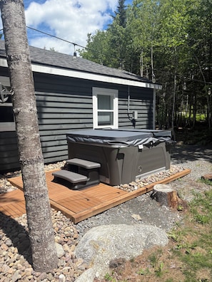 A hot tub with a wooden deck, surrounded by a stone border, next to a house with a window and a chimney.