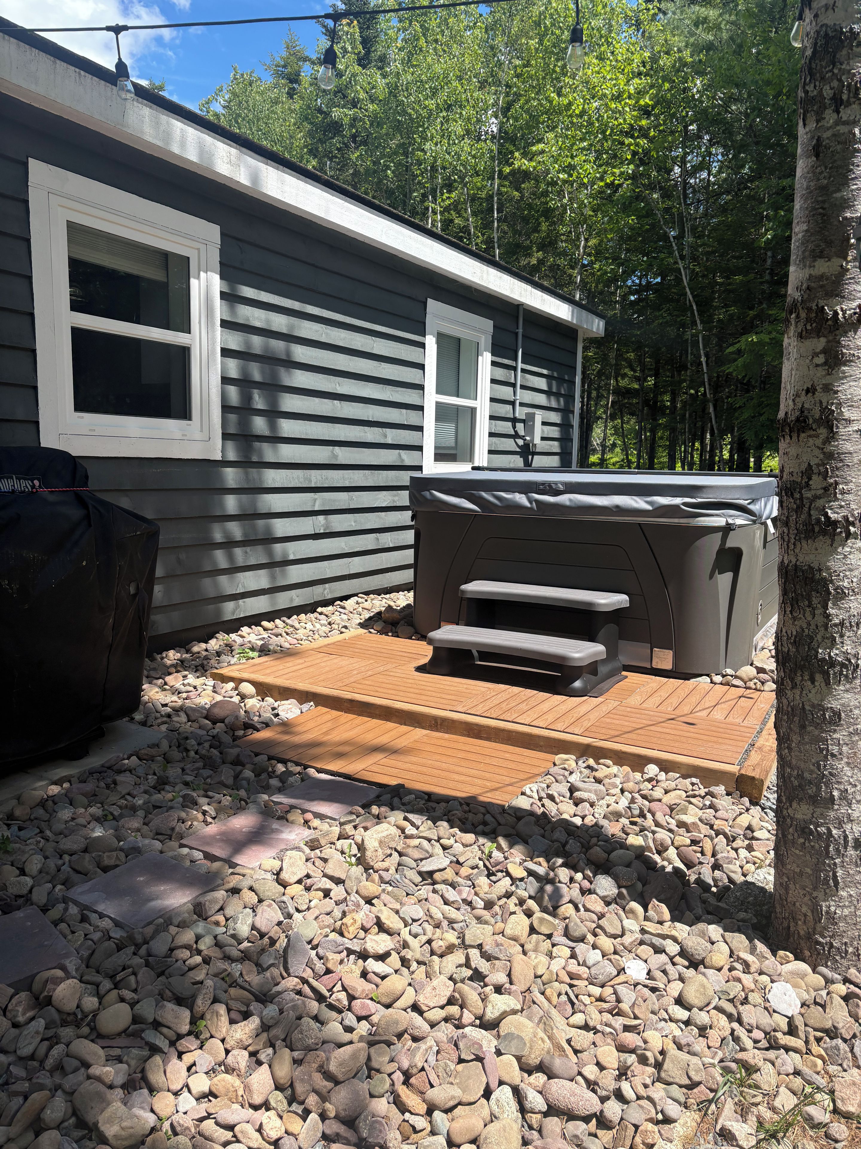 A patio area with a hot tub, a barbecue grill, and a wooden deck surrounded by rocks.