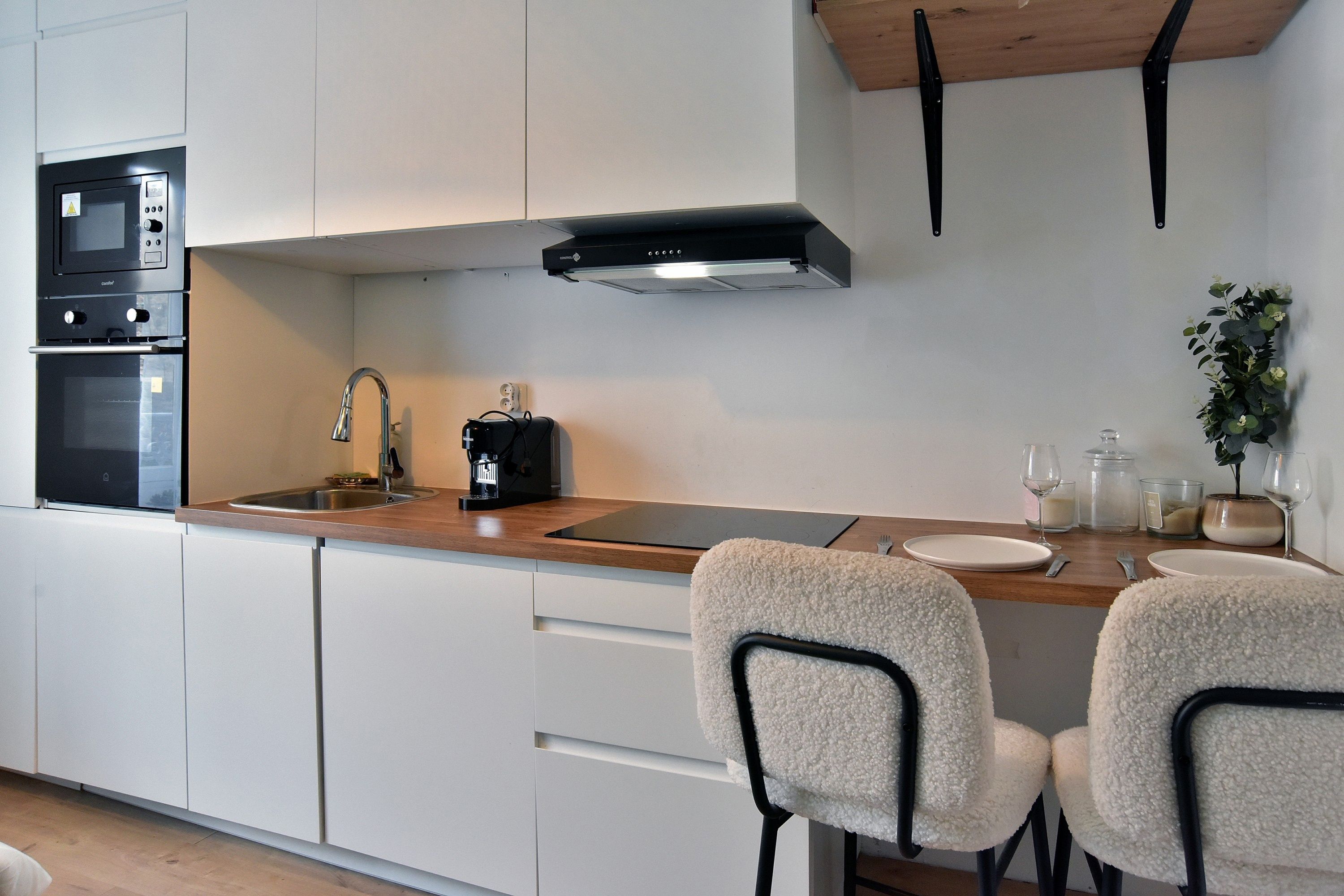 Una cocina moderna con muebles blancos, un lavamanos de acero inoxidable, una máquina de café y dos sillas blancas con estructura negra.