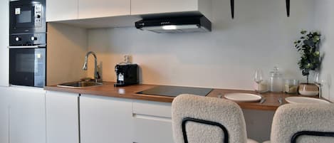 Une cuisine moderne avec des meubles blancs, un évier en acier inoxydable, une machine à café et deux chaises blanches à la structure noire.
