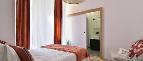 Un dormitorio con una cama, una silla, una lámpara colgante y vista a otra habitación.