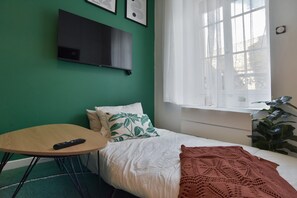 Ein Zimmer mit einem Bett, einem an der Wand befestigten Fernseher, einem Fenster mit Vorhängen und einer Pflanze.