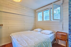A bedroom with a bed, a wooden nightstand, and a window with a view of greenery. - Flore - au cœur du bassin d'Arcachon (Arcachon)
