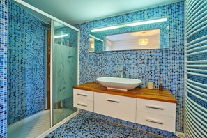 A bathroom with a glass shower enclosure, a white sink with a wooden countertop, and walls covered with blue mosaic tiles. - Flore - au cœur du bassin d'Arcachon (Arcachon)