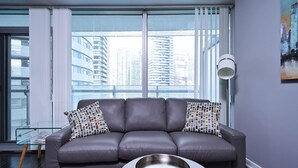 Una sala moderna con un sofá de cuero gris, una mesa de centro de vidrio y vistas de edificios altos a través de grandes ventanales.