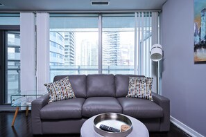 Ein modernes Wohnzimmer mit einem grauen Ledersofa, einem Glastisch und Blick auf hohe Gebäude durch große Fenster.