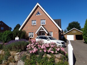 Ein zweistöckiges Backsteinhaus mit einem Auto auf der Auffahrt, umgeben von blühenden Pflanzen und einer Steinmauer.