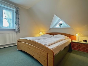 Ein Schlafzimmer mit einem hölzernen Bett, einem kleinen Nachttisch, einem Fenster mit Vorhängen und einem Oberlicht.