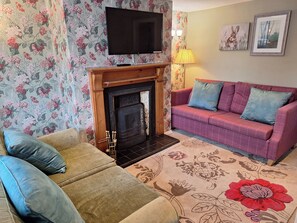 A living room with a fireplace, a sofa, a television, and floral wallpaper. - Fryers Cottage (Scarborough)