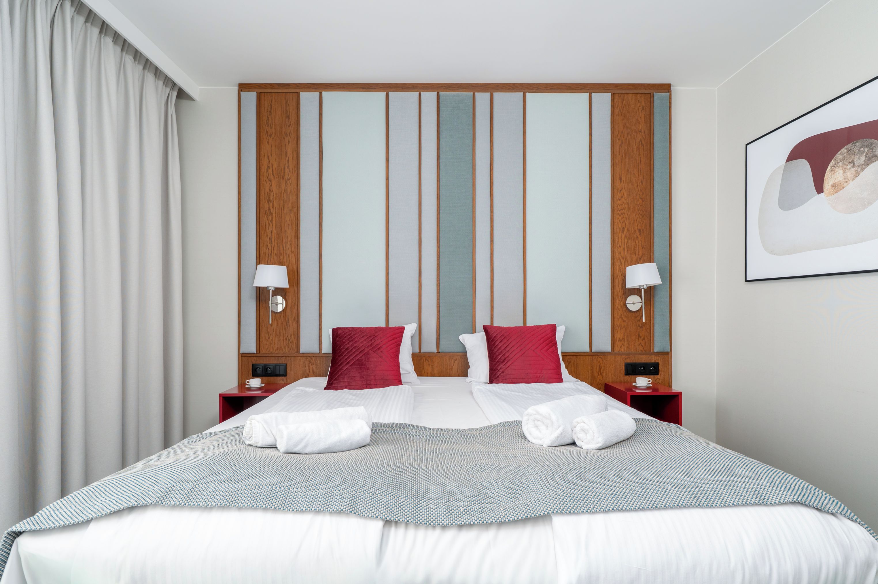 Une chambre d’hôtel avec un grand lit, deux oreillers rouges, une tête de lit en bois, une table de chevet avec une lampe et un tableau encadré au mur.