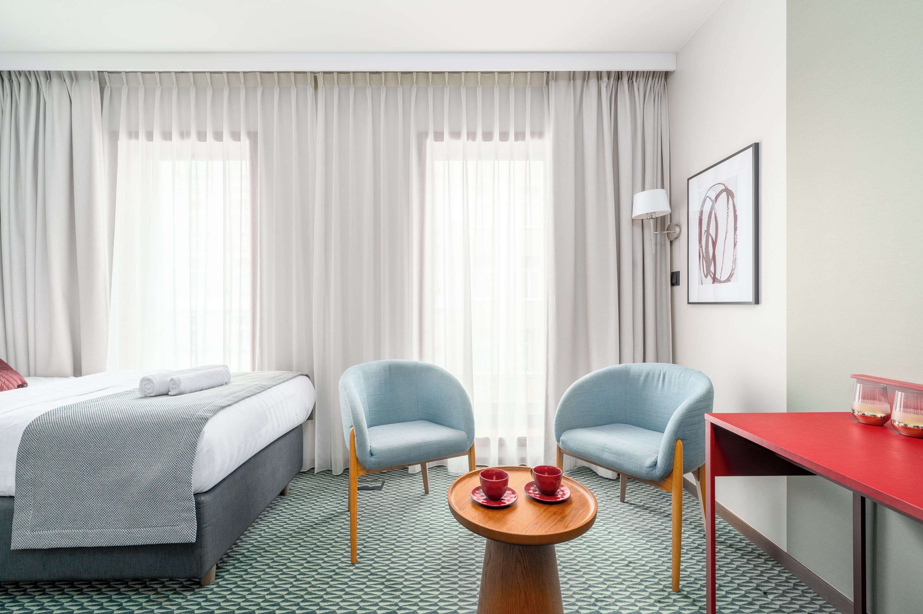A hotel room with a bed, two chairs, a small table with cups, a lamp, and a painting on the wall.