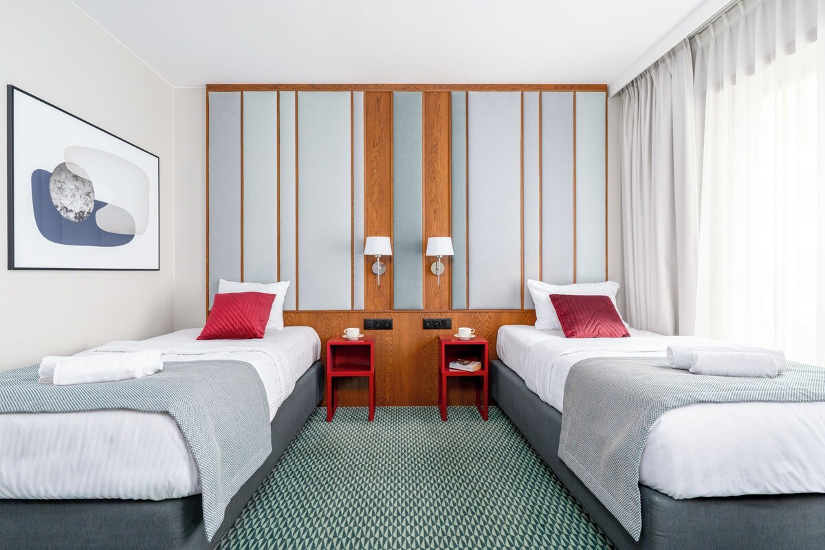 Een hotelkamer met twee bedden, een wandpaneel van hout, een ingelijst schilderij, twee bedlampen en uitzicht op de omgeving.