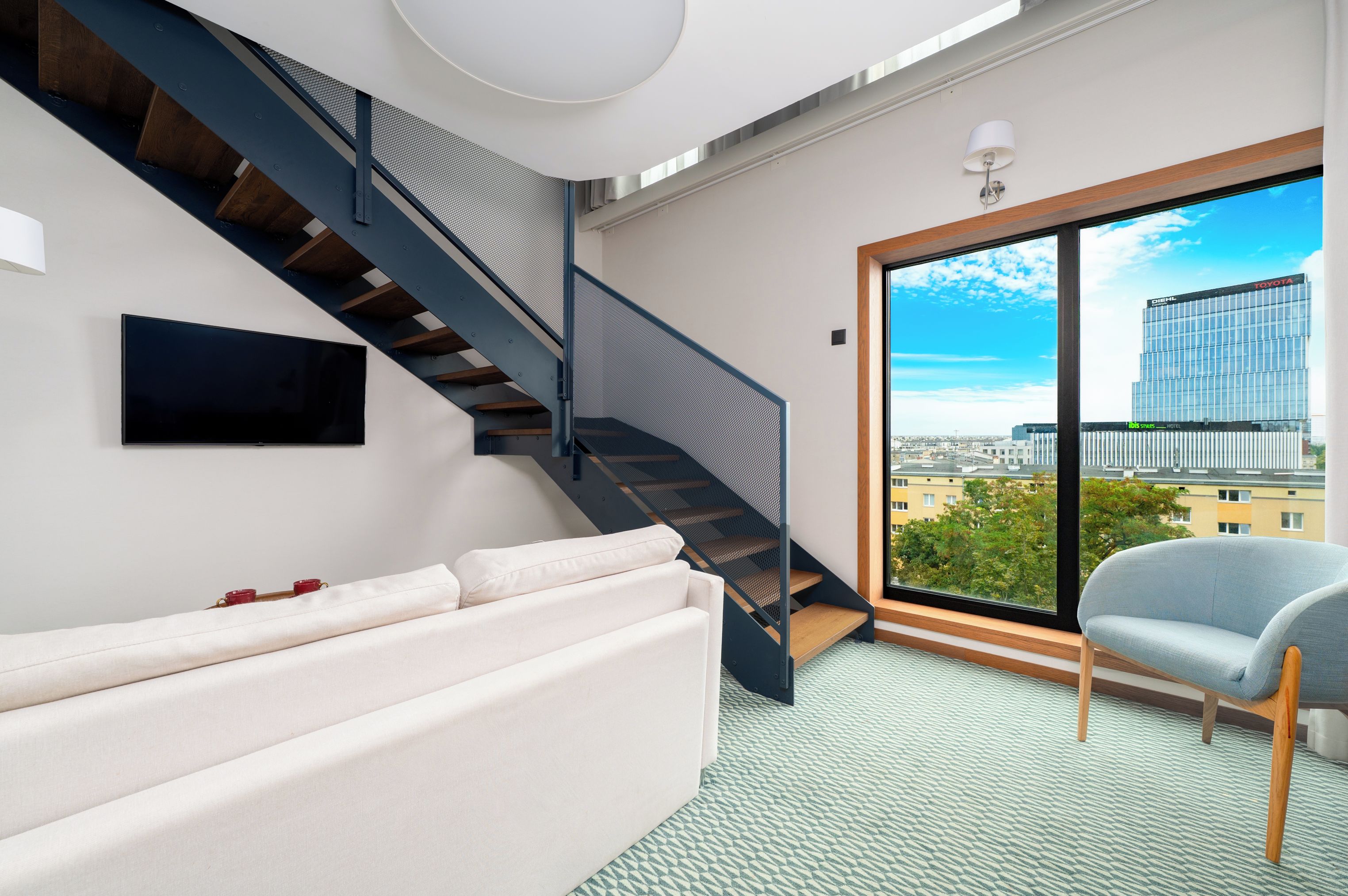 Una sala de estar moderna con una escalera, un sofá, una silla y un ventanal con vistas a la ciudad.