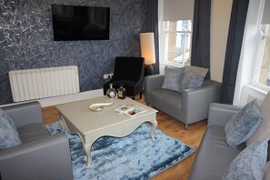 A modern living room with a flat-screen TV, a black armchair, grey sofas, a white coffee table with magazines, and a blue patterned rug. - Cornerhouse Apartment 2 (Llandudno)