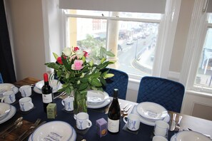A dining table set with plates, cups, and a bottle of wine, with a view of a street through the window. - Cornerhouse Apartment 2 (Llandudno)