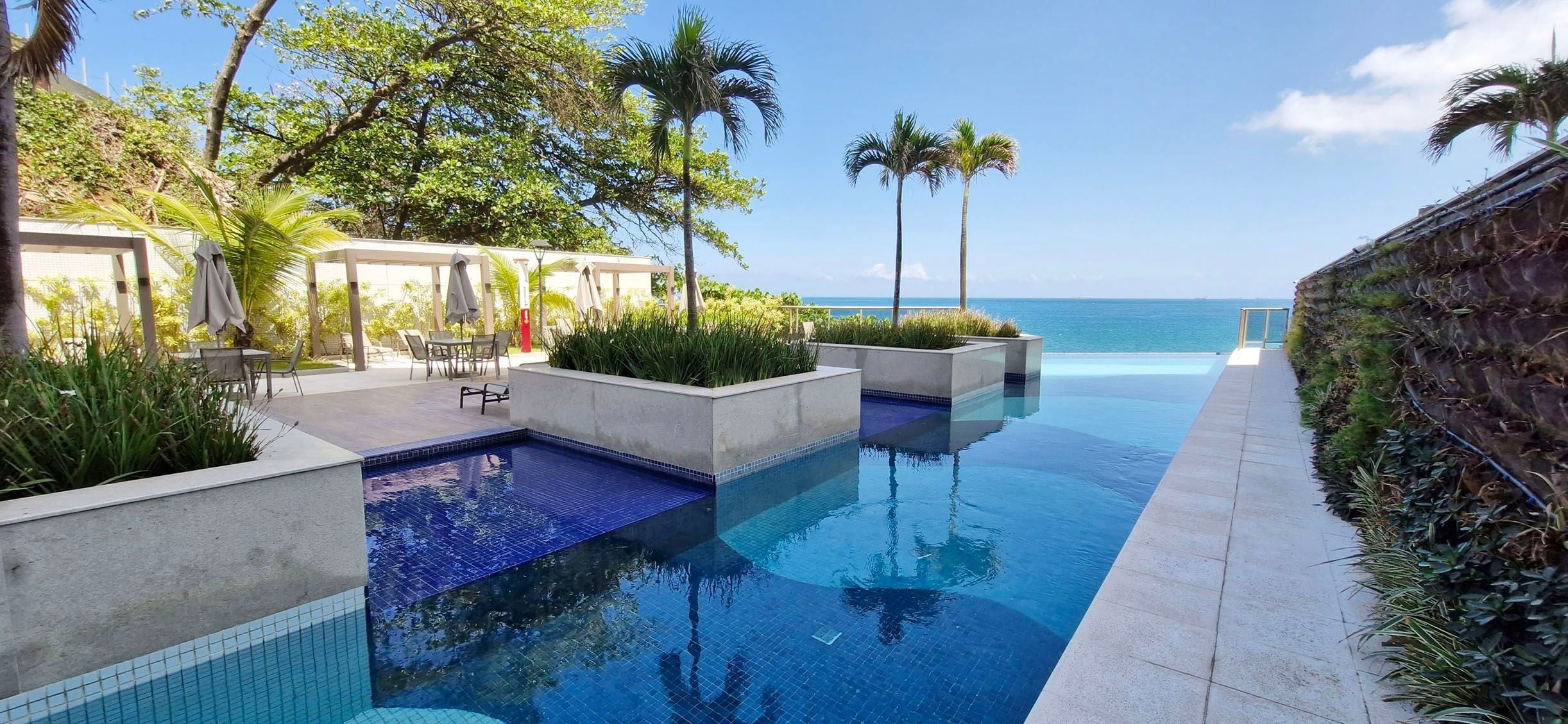 Área da piscina com vista para o oceano, rodeada por palmeiras e mobiliário de exterior.