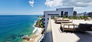A rooftop terrace with a view of the ocean, outdoor furniture, and a modern building. - COOND0126 - Beach Class Salvador (Salvador)