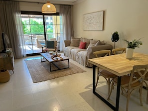 Ein Wohnzimmer mit einer Couch, einem Couchtisch, einem Esstisch mit Stühlen, einem großen Fenster mit Vorhängen und einer Hängelampe.