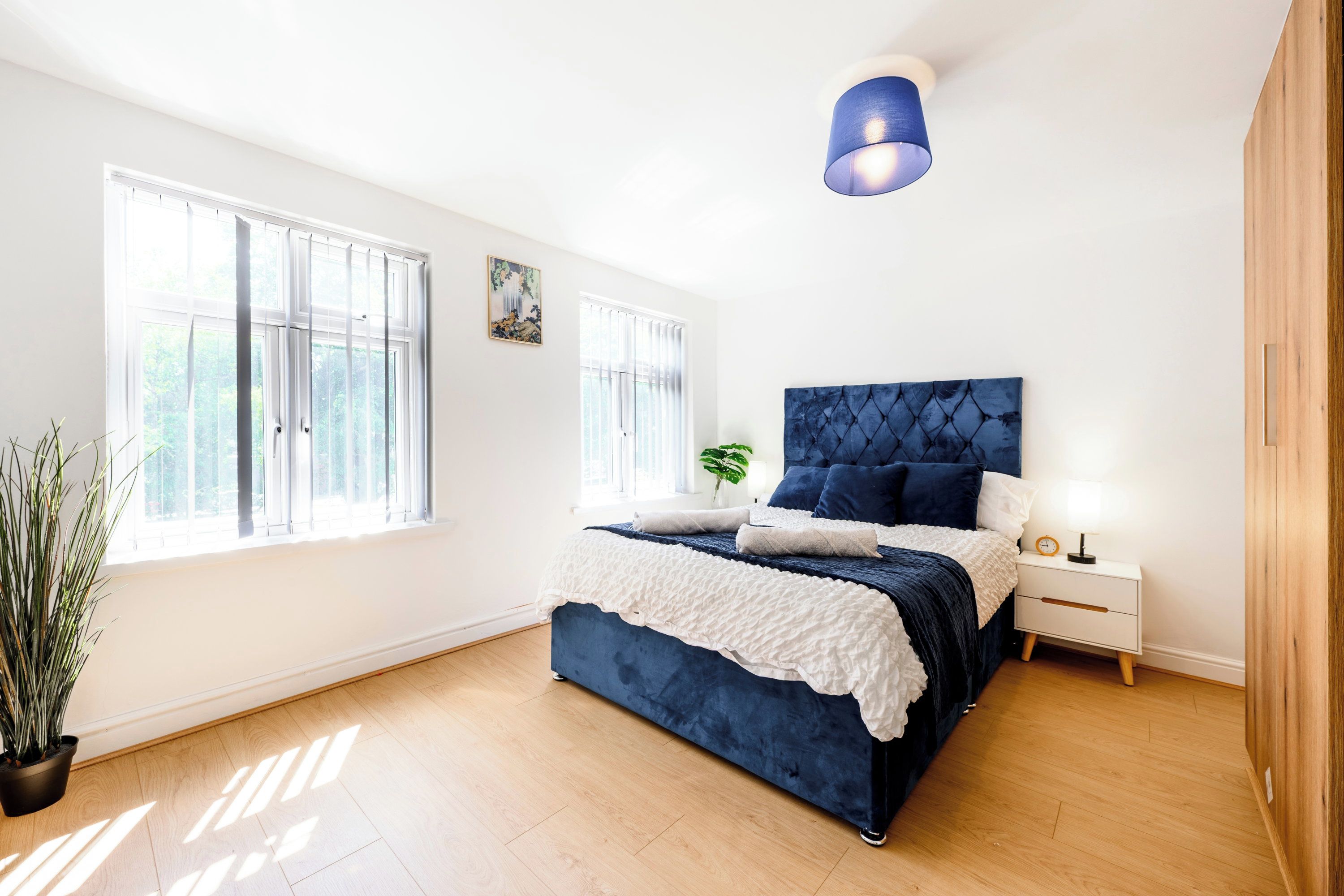 A bedroom with a large bed, a nightstand, a potted plant, and a window with blinds.