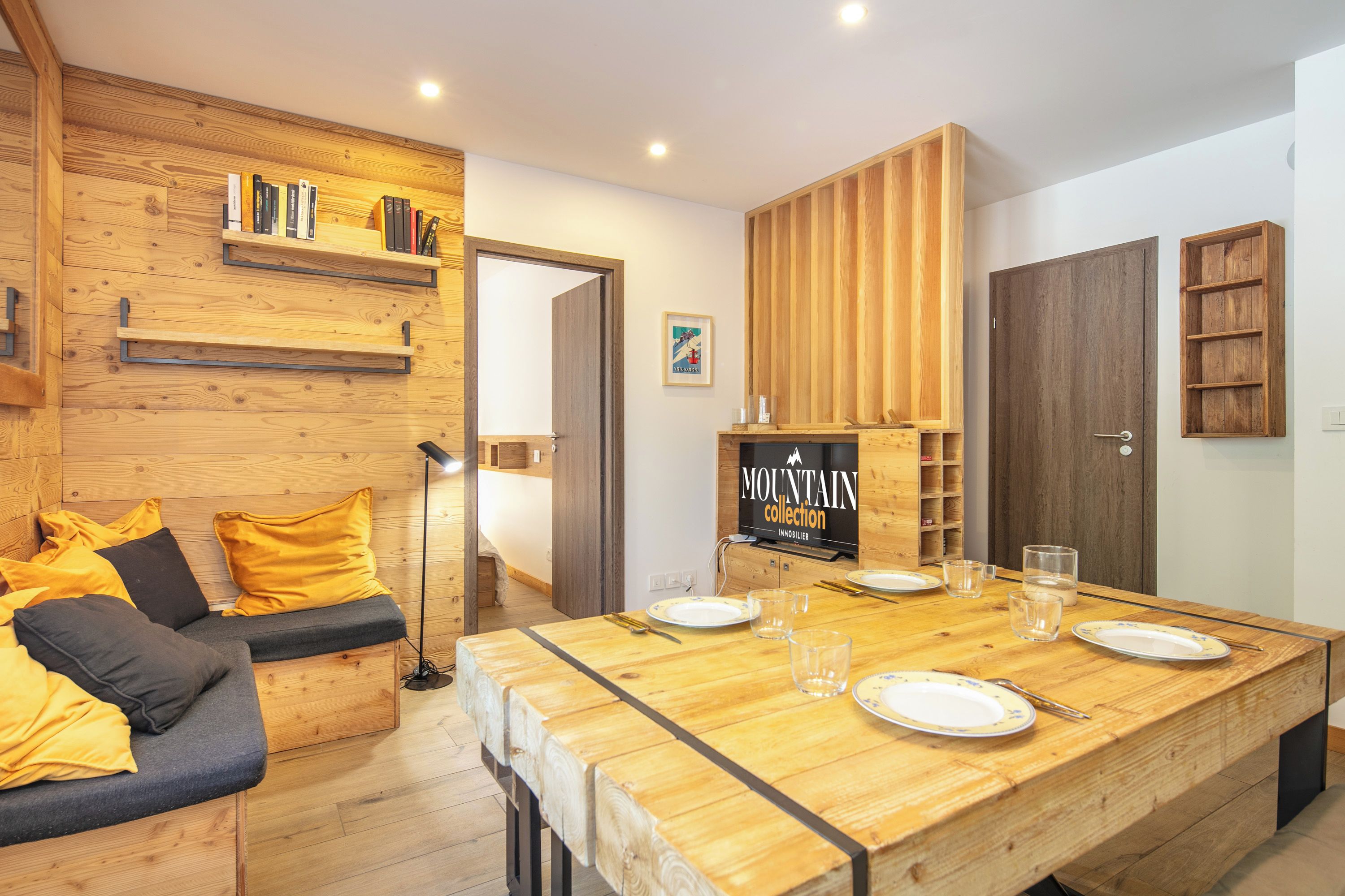 Un intérieur moderne avec une table à manger en bois, un canapé avec des coussins et un téléviseur fixé au mur qui diffuse la collection « MOUNTAIN ».