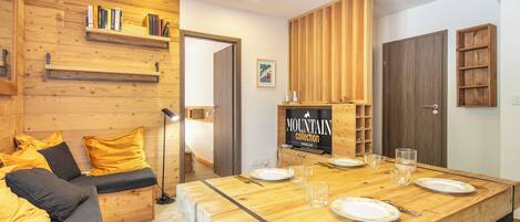 Un intérieur moderne avec une table à manger en bois, un canapé avec des coussins et un téléviseur fixé au mur qui diffuse la collection « MOUNTAIN ».