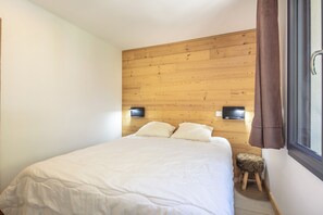 Een slaapkamer met een houten hoofdbord, een bed met wit beddengoed, een klein krukje en een raam met gordijnen.