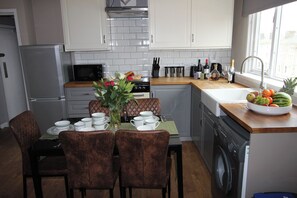 A modern kitchen with white cabinetry, a stainless steel refrigerator, a microwave, and a dishwasher. There is a dining table with chairs and a vase of flowers. - Cornerhouse Apartment 4 (Llandudno)