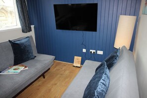 A room with a grey sofa, a blue wall, a mounted TV, and a lamp. - Cornerhouse Apartment 4 (Llandudno)