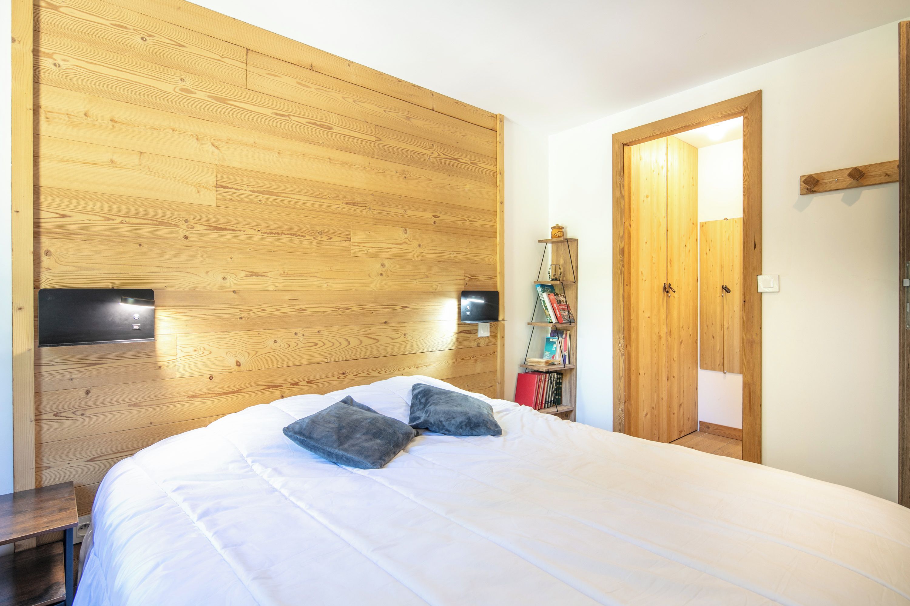 Une chambre à coucher avec une tête de lit en bois, un lit avec une literie blanche et deux oreillers, une table de chevet et une armoire en bois dotée d’un miroir.