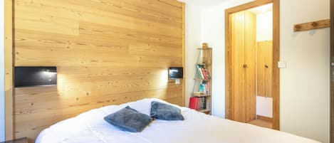 Une chambre à coucher avec une tête de lit en bois, un lit avec une literie blanche et deux oreillers, une table de chevet et une armoire en bois dotée d’un miroir.