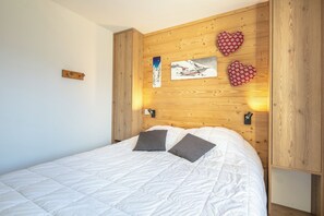 A bedroom with a wooden headboard, a bed with white bedding, two pillows, and wall-mounted artwork. - travelski home premium - Résidence La Mariande - 3 Pièces 6 personnes N°3.3 (Les Deux Alpes)