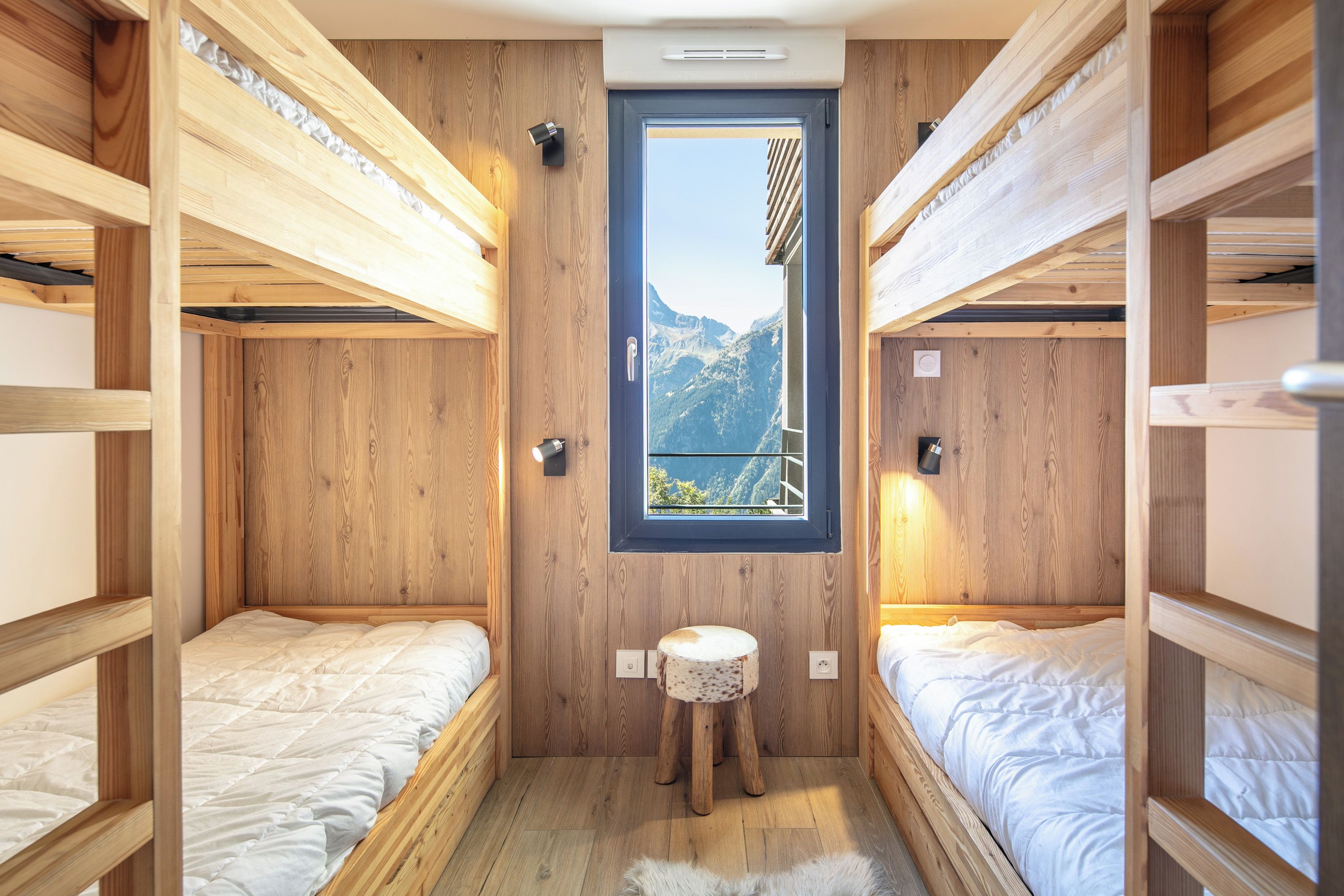 Ein kleines Zimmer mit zwei Etagenbetten, einem kleinen Hocker und einem Fenster mit Bergblick.