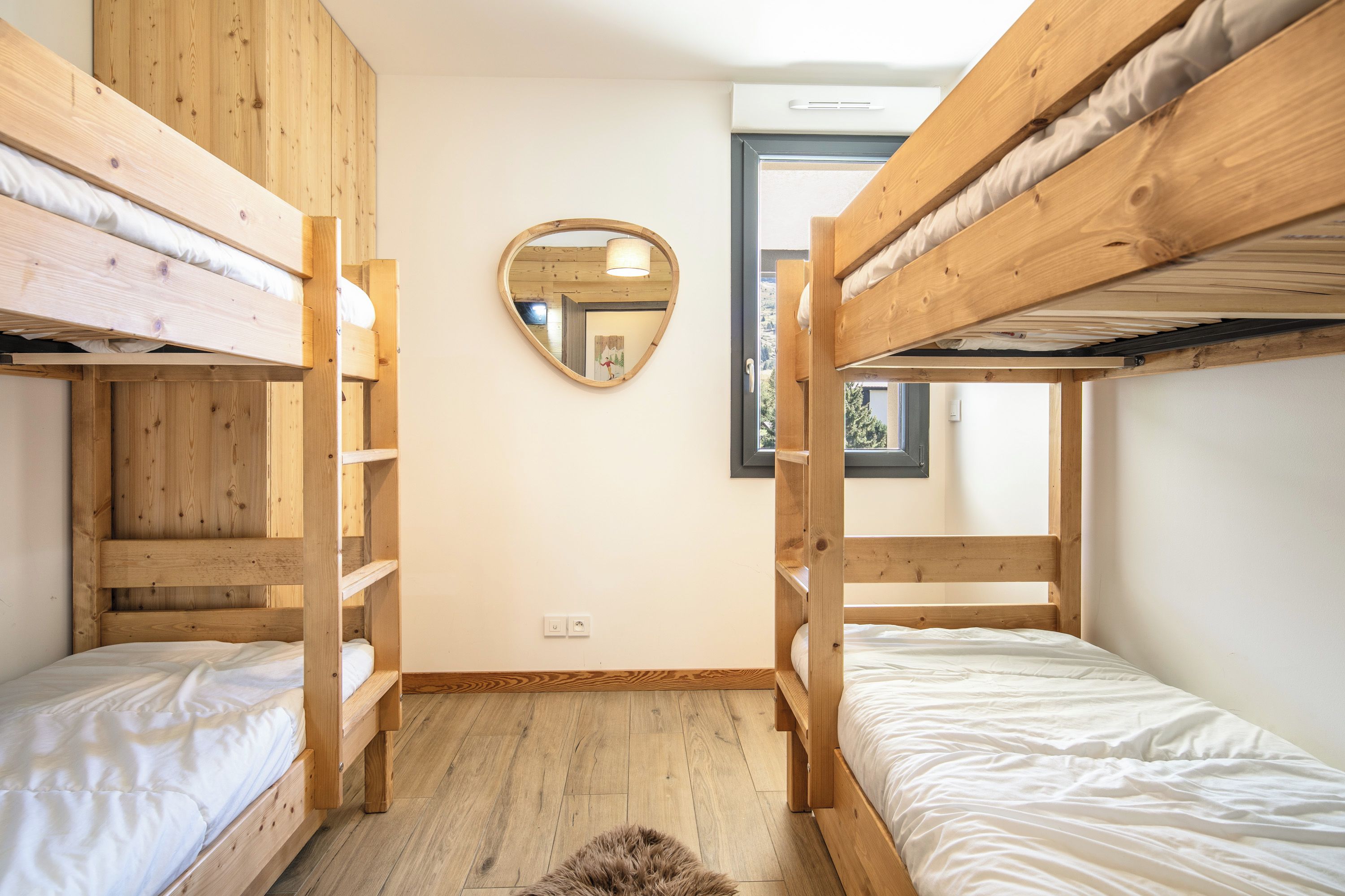 A bunk bed room with a wooden floor, a round mirror on the wall, and a window with a view.