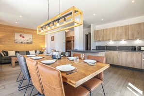 A modern kitchen with wooden cabinets, a dining area with a wooden table set for four, and a living area with a sofa and a painting on the wall. - travelski home premium - Résidence La Mariande - 4 Pièces 8 personnes N°1.2 (Les Deux Alpes)