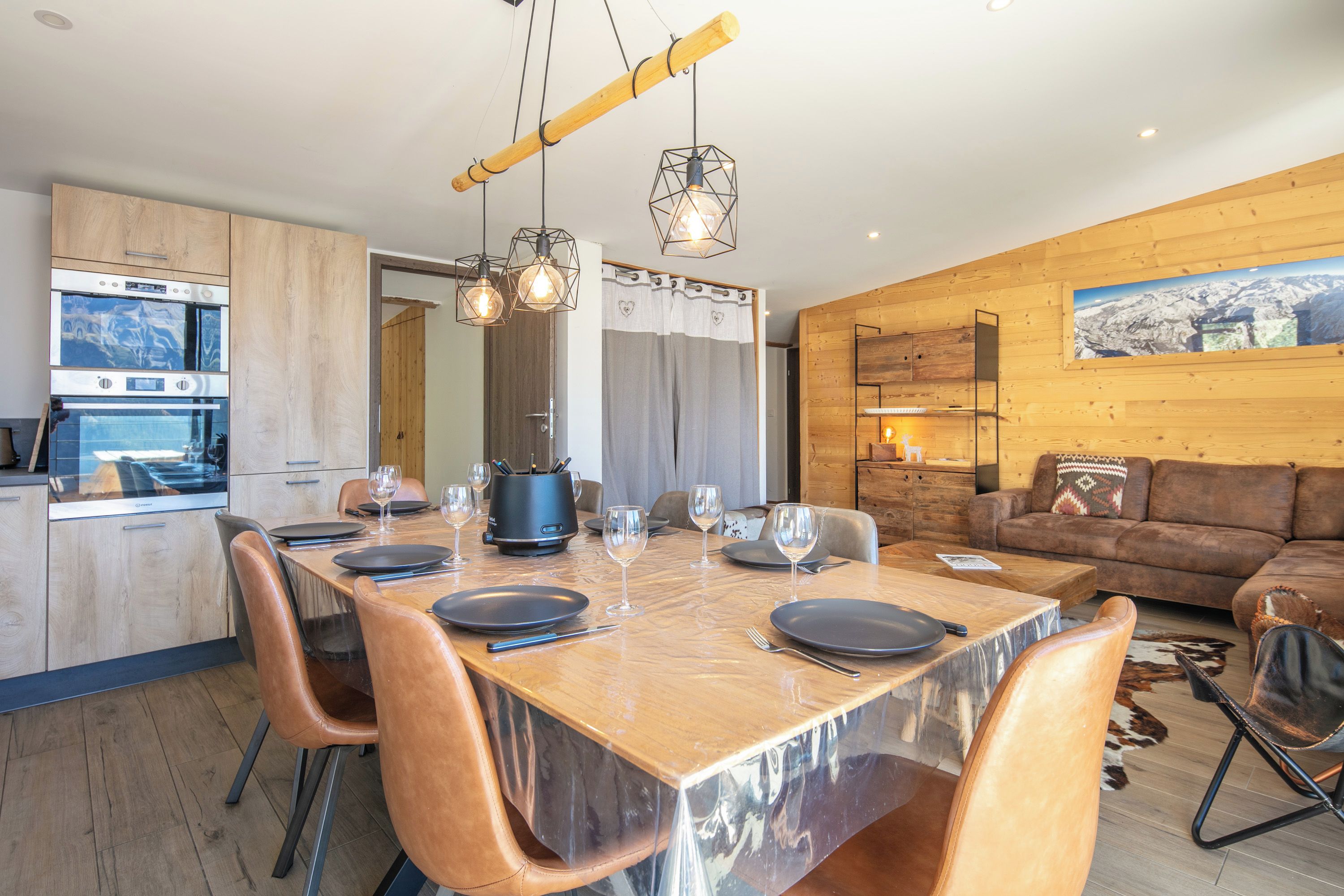 A modern kitchen with built-in appliances, a dining area with a wooden table and chairs, and a living room with a sofa and a mounted picture.