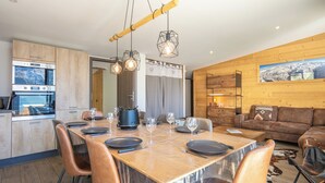 Cozinha moderna com eletrodomésticos embutidos, sala de jantar com mesa e cadeiras de madeira, e sala de estar com sofá e quadro pendurado.