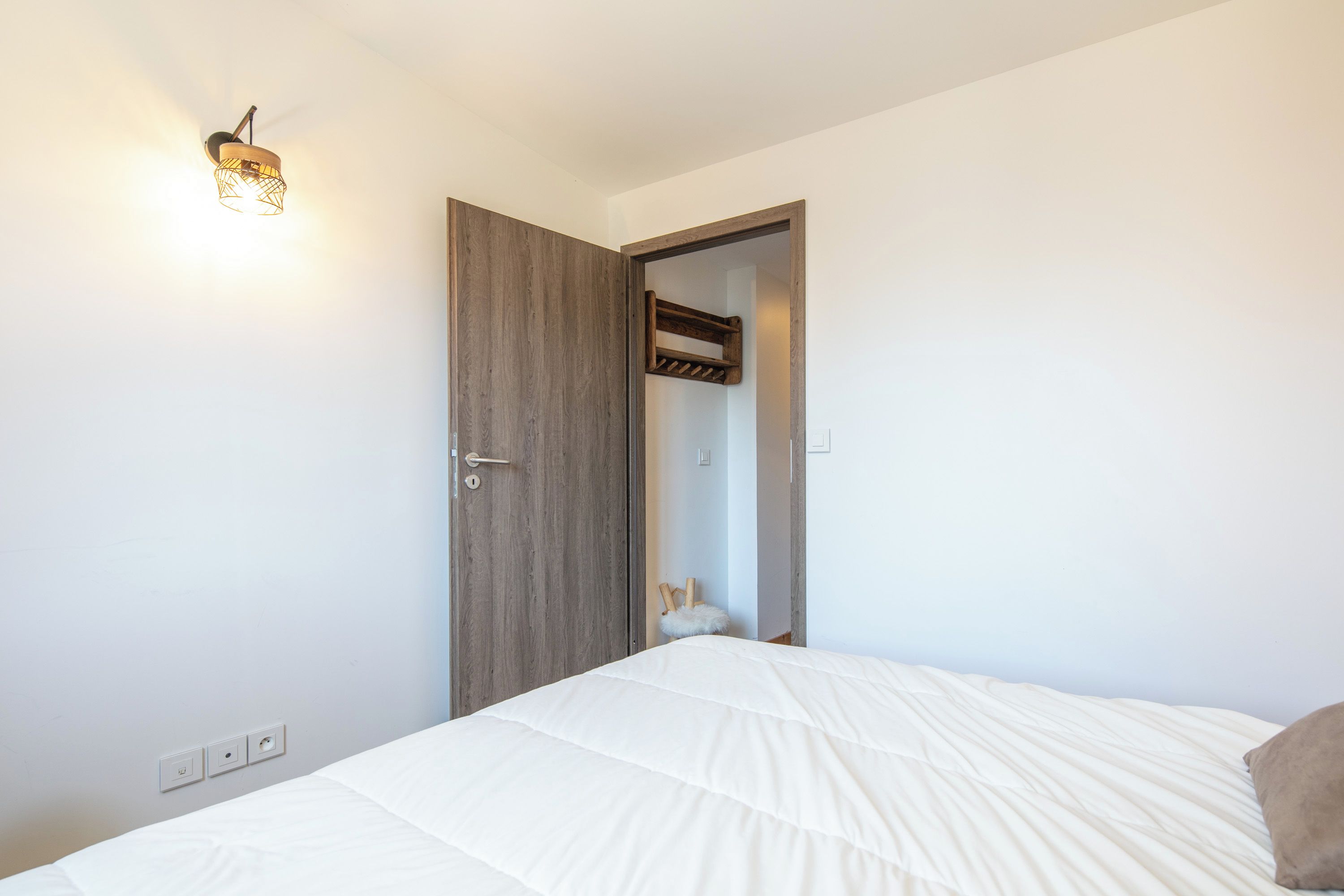 Quarto com cama, porta de madeira e luminária fixada na parede.